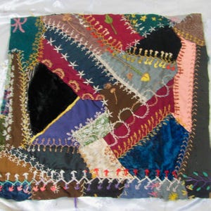 May include: A square patchwork textile piece featuring an array of multi-colored fabric scraps in shades of deep blue, burgundy, black, and brown. The segments are joined by decorative white and yellow embroidery stitches in various patterns.