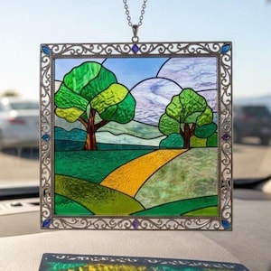 Könnte beinhalten: Buntglasfenster mit einer Landschaftsszene mit Bäumen, einem Weg und Hügeln. Das Kunstwerk ist in einem dekorativen, silberfarbenen Metallrahmen mit blauen und violetten Akzenten eingefasst. Das Panel wird von einer silbernen Kette gehalten.