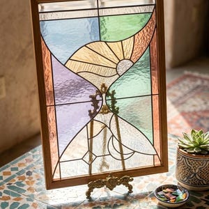 May include: A rectangular stained glass panel with a wooden frame, featuring a sunburst design in shades of yellow, orange, blue, and purple. The panel is displayed on a decorative stand. The artwork is a decorative home decor item.