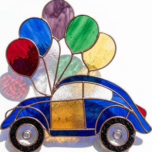 May include: A stained glass suncatcher featuring a blue car with a yellow door and textured white windows. Attached to the roof are five colourful glass balloons in red, blue, purple, green, and yellow, all held together by copper foil lines.