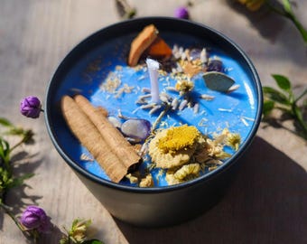 May include: A blue candle in a black container with a white wick, decorated with cinnamon sticks, dried flowers, and crystals. The text "The Eye of Venus Metaphysical Shop" is at the top of the image. The candle sits on a wooden surface.