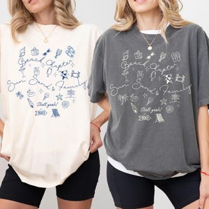 May include: Two oversized t-shirts, one cream and one charcoal, each with a beach-themed graphic. The design includes the words "Beach Chapter, Sun, Sand & Family" with illustrations of beach items like sunglasses, ice cream, and seashells. The phrase "Shell yeah!" is also included.