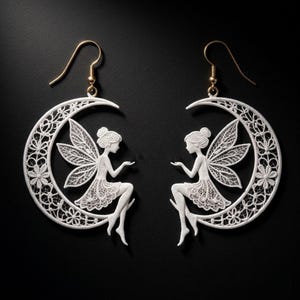 Puede incluir: Pendientes de hadas blancos con ganchos dorados. Cada pendiente presenta una figura de hada sentada en una luna creciente. La luna y el hada están intrincadamente detalladas con patrones florales y de encaje, sobre un fondo negro.