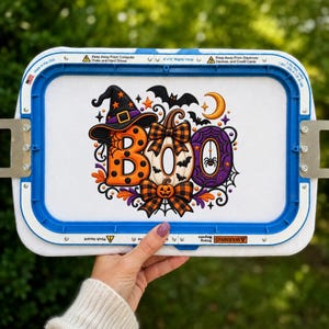 May include: An 8x13 inch blue embroidery hoop containing white fabric with a colorful Halloween-themed machine embroidery design. The design features the word BOO in orange, white, and purple, decorated with a witch hat, bows, and bats.