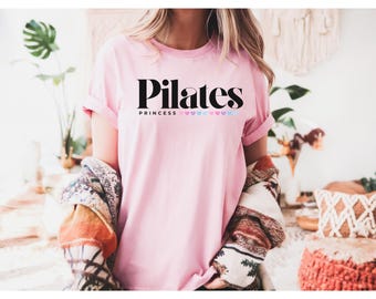 Puede incluir: Camiseta rosa claro con la frase "Pilates Princess" en negro. La palabra "Pilates" está encima de "Princess", con una fila de corazones rosas y azules debajo. Las mangas están dobladas.
