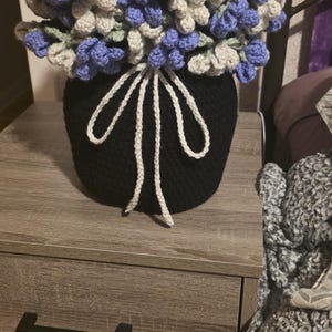 May include: A handmade crochet flower arrangement in a black pot. The flowers are a mix of blue, gray, and green, with a white decorative tie. The arrangement sits on a wooden nightstand.