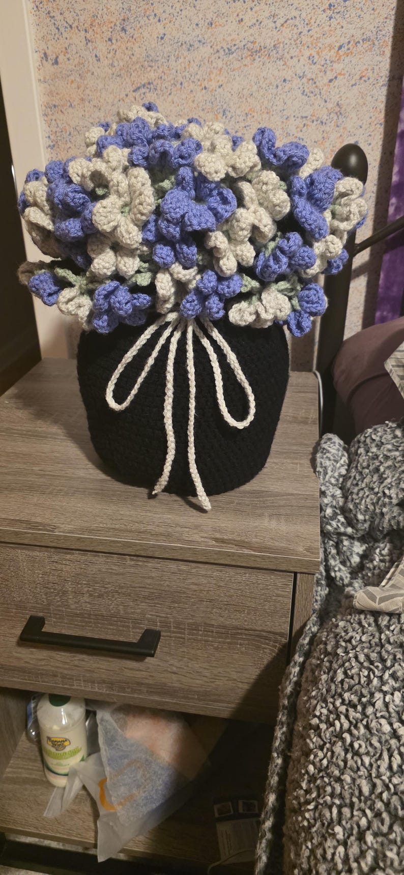 May include: A handmade crochet flower arrangement in a black pot. The flowers are a mix of blue, gray, and green, with a white decorative tie. The arrangement sits on a wooden nightstand.