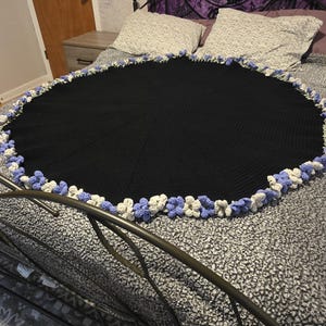 May include: A black, round crocheted blanket with a decorative border of white and light blue crocheted flowers. The blanket is displayed on a bed with a patterned gray and white bedspread.