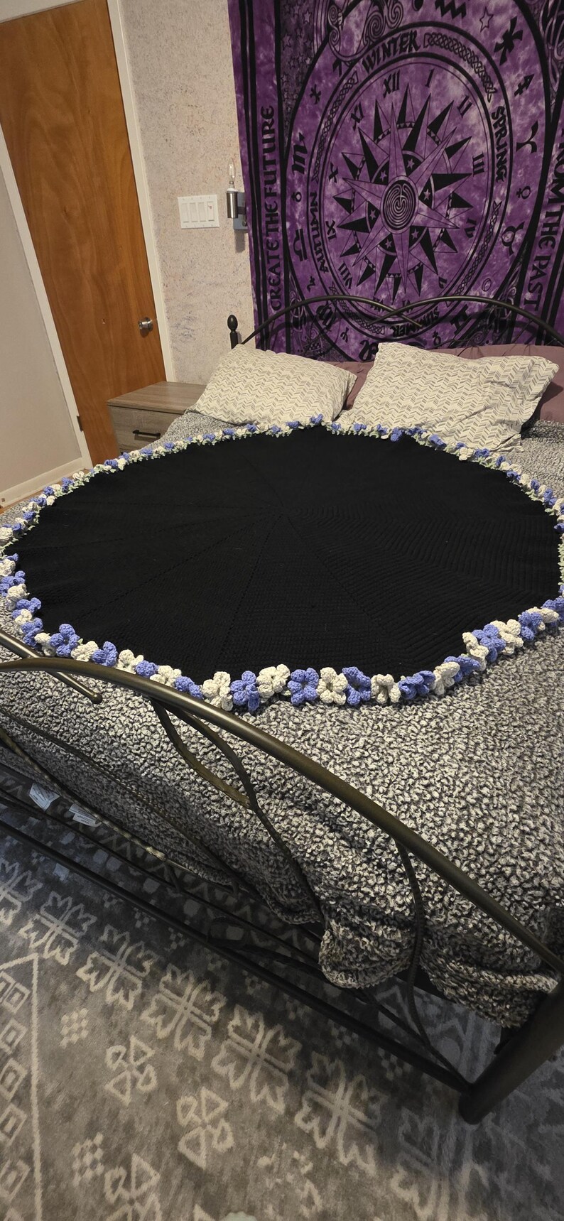 May include: A black, round crocheted blanket with a decorative border of white and light blue crocheted flowers. The blanket is displayed on a bed with a patterned gray and white bedspread.