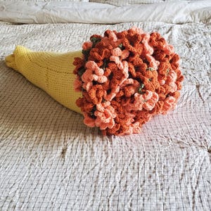 May include: A handmade crochet bouquet in a yellow cone-shaped holder. The bouquet features orange and peach-colored flowers with green accents. The arrangement is placed on a checkered bedspread.