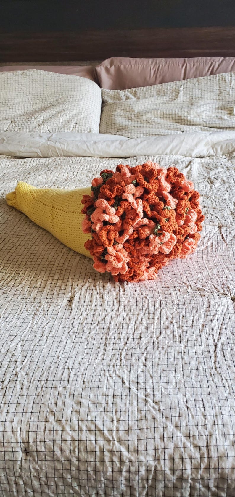 May include: A handmade crochet bouquet in a yellow cone-shaped holder. The bouquet features orange and peach-colored flowers with green accents. The arrangement is placed on a checkered bedspread.