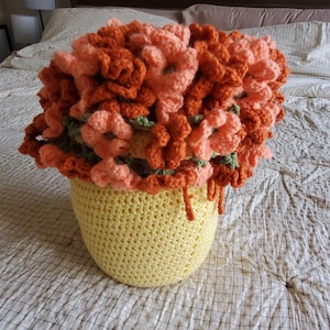May include: A handmade crochet flower arrangement in a yellow pot. The flowers are a mix of orange and peach, with green accents. The pot is a light yellow color. The arrangement sits on a bed with a white and blue checkered pattern.