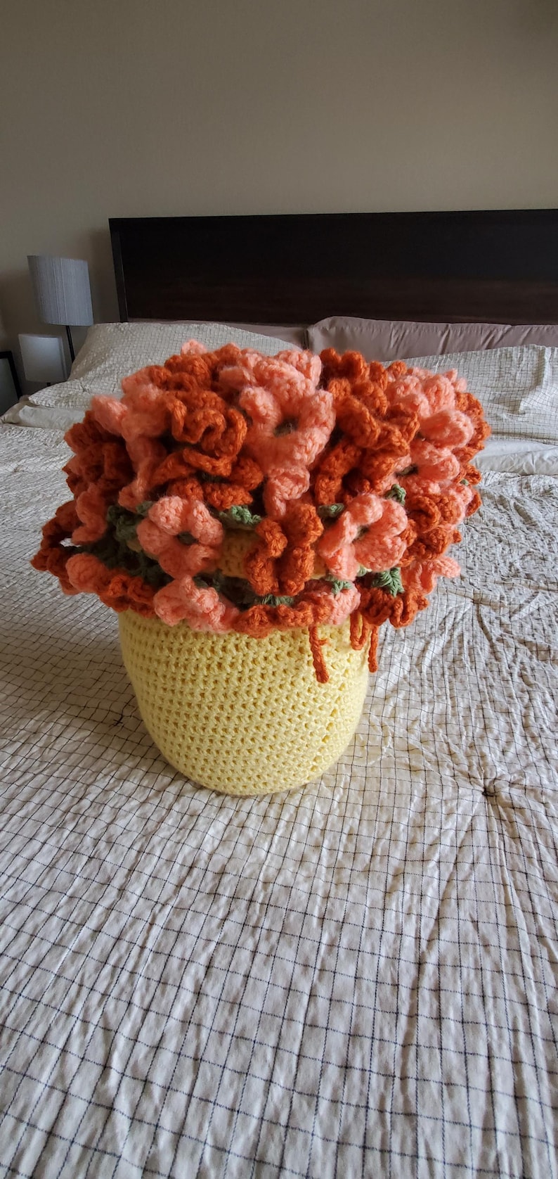 May include: A handmade crochet flower arrangement in a yellow pot. The flowers are a mix of orange and peach, with green accents. The pot is a light yellow color. The arrangement sits on a bed with a white and blue checkered pattern.