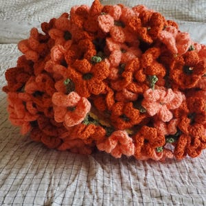 May include: A handmade crochet floral pillow in shades of orange and peach. The pillow is made of many individual crochet flowers with green centers, arranged to create a dense, textured surface. The pillow is resting on a white and gray checkered bedspread.