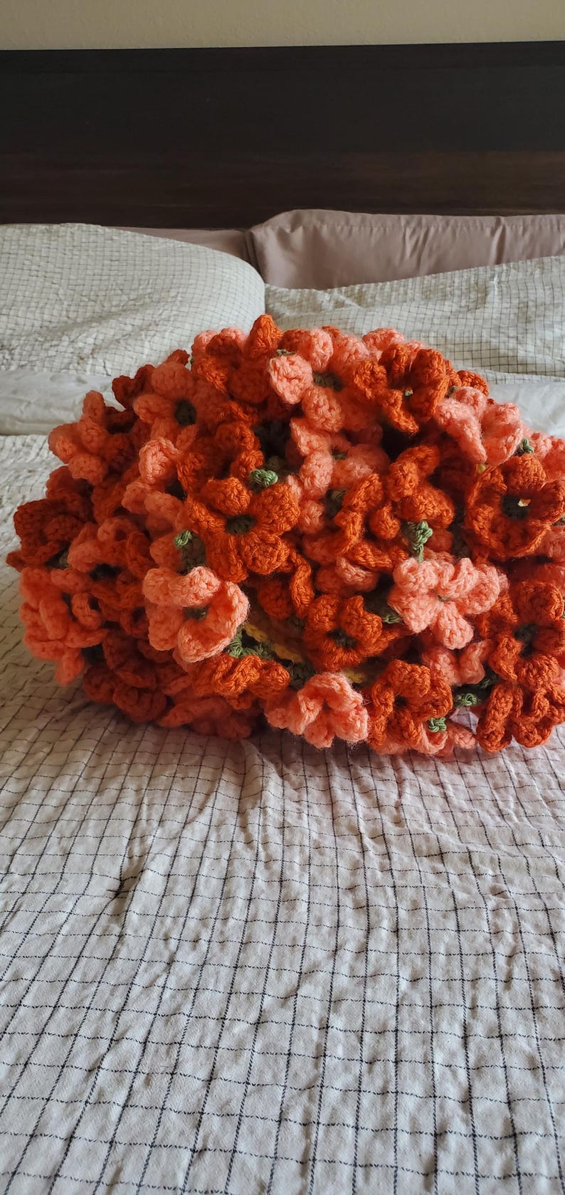 May include: A handmade crochet floral pillow in shades of orange and peach. The pillow is made of many individual crochet flowers with green centers, arranged to create a dense, textured surface. The pillow is resting on a white and gray checkered bedspread.