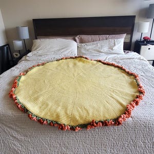 May include: A round, yellow crocheted blanket with a decorative orange and green trim. The blanket is placed on a bed with a neutral-colored comforter. The blanket's design features a central starburst pattern.