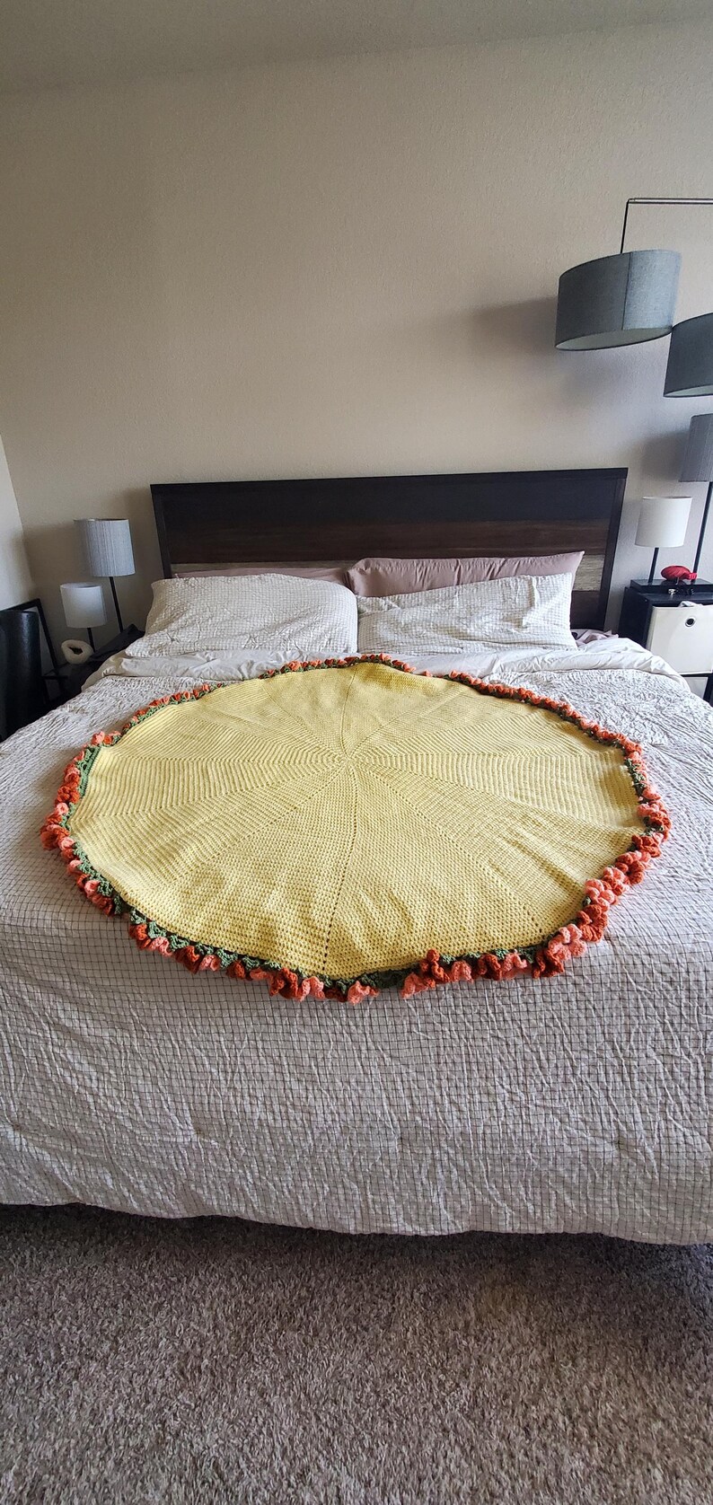 May include: A round, yellow crocheted blanket with a decorative orange and green trim. The blanket is placed on a bed with a neutral-colored comforter. The blanket's design features a central starburst pattern.