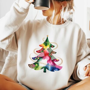 May include: White sweatshirt featuring a colorful Christmas tree design. The tree is composed of various patterns and colors, including red gingham, leopard print, and green plaid. A gold star tops the tree, with gold string lights.