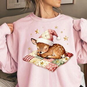 May include: Pale pink sweatshirt with a watercolour illustration of a sleeping fawn wearing a Christmas hat. The fawn rests on a patchwork pillow with gold star accents. A festive and cosy design.