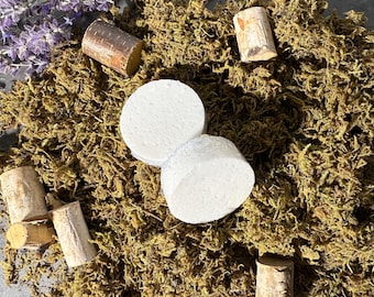 May include: Two white, round bath bombs and several cylindrical corks are arranged on a bed of green moss. A sprig of purple lavender adds a touch of color. The natural elements create a spa-like aesthetic.