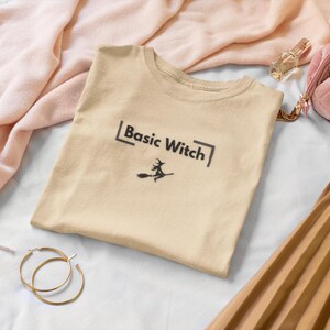 May include: A beige t-shirt with the text "Basic Witch" and a witch on a broomstick graphic. The shirt is surrounded by a pink scarf, gold hoop earrings, a pink quilted bag, and a pleated skirt.