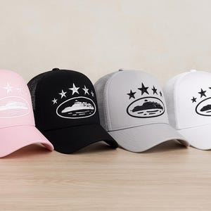 Peut inclure: Quatre casquettes de camionneur roses, noires, grises et blanches. Chaque casquette présente un motif ovale blanc avec un bateau et des étoiles. La casquette rose a un dos en filet rose, tandis que la casquette noire a un dos en filet noir.