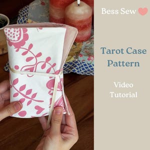 以下が含まれることがあります： ピンクの花柄とクリーム色のリボン留めが付いた白いタロットカードケース。ケースは開いており、ピンク色の内側が見えます。画像には「Bess Sew」と「Tarot Case Pattern Video Tutorial」という文字が含まれています。