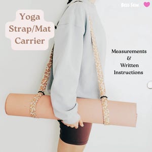 May include: A yoga mat carrier with a floral patterned strap. The carrier holds a rolled-up pink yoga mat. The text "Yoga Strap/Mat Carrier" is visible. The carrier is designed to be worn over the shoulder.
