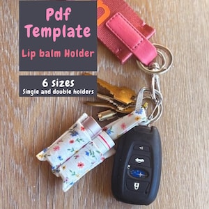 Puede incluir: Una plantilla PDF para un soporte de bálsamo labial, con el texto "Pdf Template Lip balm Holder 6 sizes Single and double holders". El soporte es blanco con un estampado floral y está sujeto a un llavero con llaves y un mando de coche.