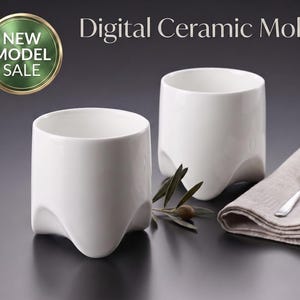 Puede incluir: Dos tazas de cerámica blanca con acabado liso y una base de cuatro patas. El texto Digital Ceramic Mold aparece en la parte superior. Una pequeña cuchara de plata descansa sobre un paño beige doblado junto a una ramita con hojas verdes.