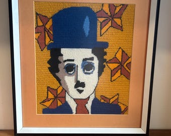 May include: A framed needlepoint artwork depicting a stylized figure wearing a blue top hat and suit with a red tie. The figure has a white face, black hair, and a mustache, set against a mustard yellow background with orange and brown star shapes.