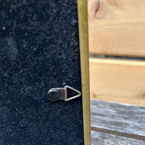 May include: A rectangular picture frame back featuring a black velvet-like surface enclosed by a thin brass-colored metal border. A small, triangular metal hanging hook is attached to the center of the black backing for wall mounting purposes.