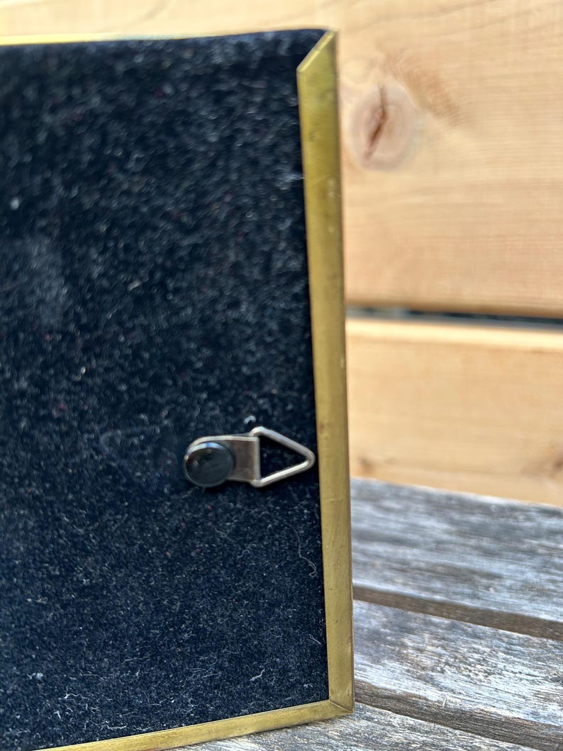 May include: A rectangular picture frame back featuring a black velvet-like surface enclosed by a thin brass-colored metal border. A small, triangular metal hanging hook is attached to the center of the black backing for wall mounting purposes.