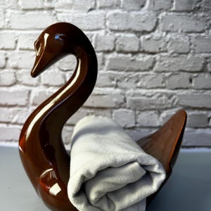 May include: A brown ceramic swan-shaped holder with a rolled-up gray towel. The swan has a glossy finish and a curved neck. The towel is made of a soft, light-colored fabric. The background is a white brick wall.