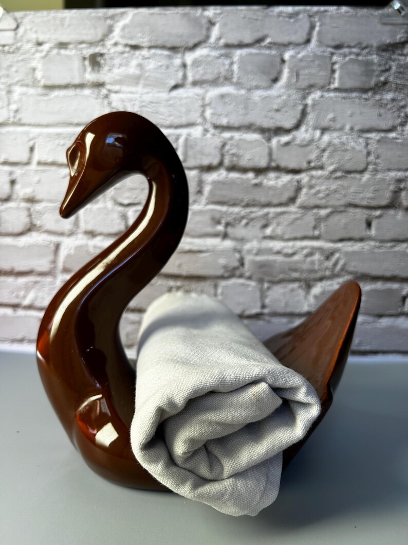 May include: A brown ceramic swan-shaped holder with a rolled-up gray towel. The swan has a glossy finish and a curved neck. The towel is made of a soft, light-colored fabric. The background is a white brick wall.