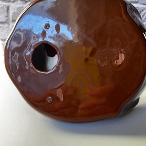 May include: A brown ceramic object with a glossy finish. The object has a rounded shape with a hole and several small imperfections. The color is a rich, warm brown, and the surface reflects light.