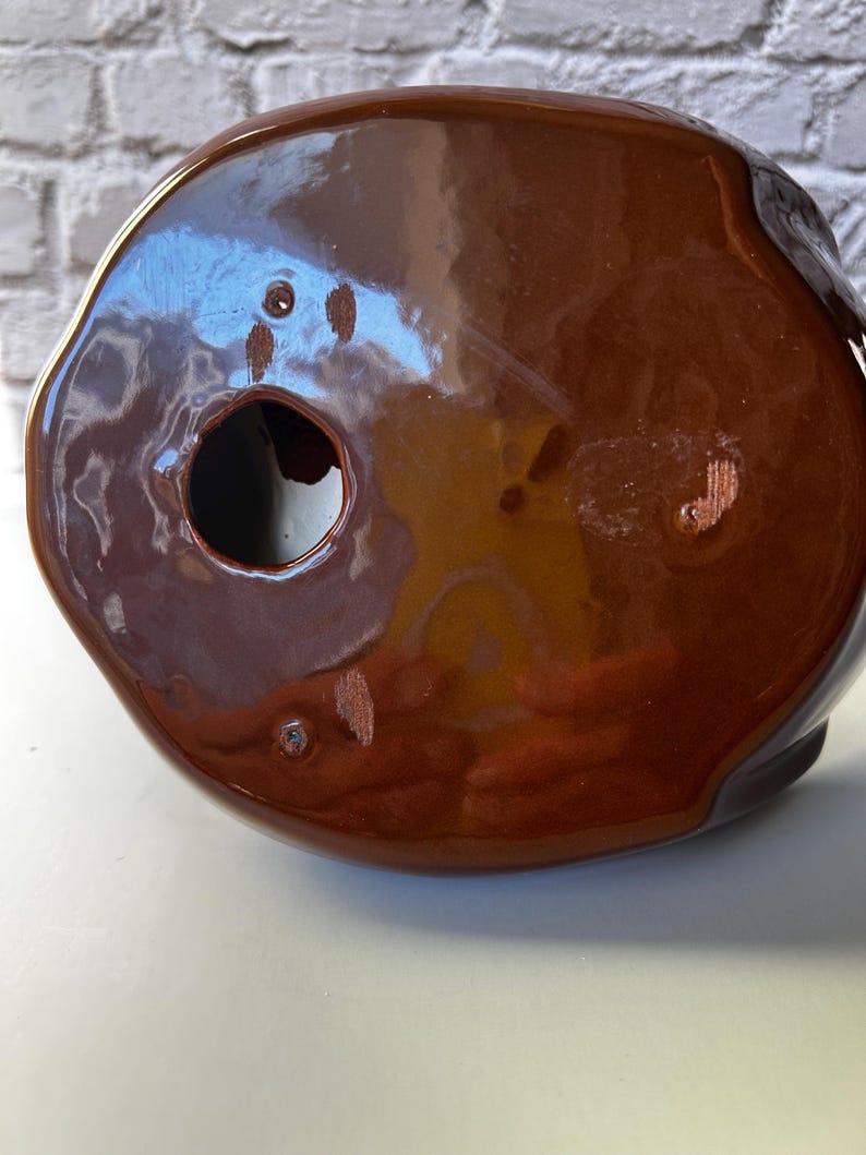 May include: A brown ceramic object with a glossy finish. The object has a rounded shape with a hole and several small imperfections. The color is a rich, warm brown, and the surface reflects light.