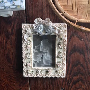 May include: A rectangular picture frame with a cream-colored border featuring sculpted roses and leaves. At the top, a sculpted bow sits above two silver-toned bells. The frame contains a black and white photograph of flowers, surrounded by a beaded edge.