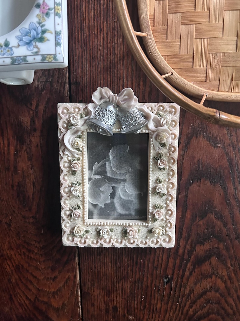 May include: A rectangular picture frame with a cream-colored border featuring sculpted roses and leaves. At the top, a sculpted bow sits above two silver-toned bells. The frame contains a black and white photograph of flowers, surrounded by a beaded edge.