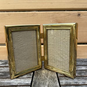May include: A double-hinged picture frame constructed from polished gold-tone metal. The frame features two rectangular openings connected by a central metal hinge, allowing it to stand upright in a folded position with light beige inserts.