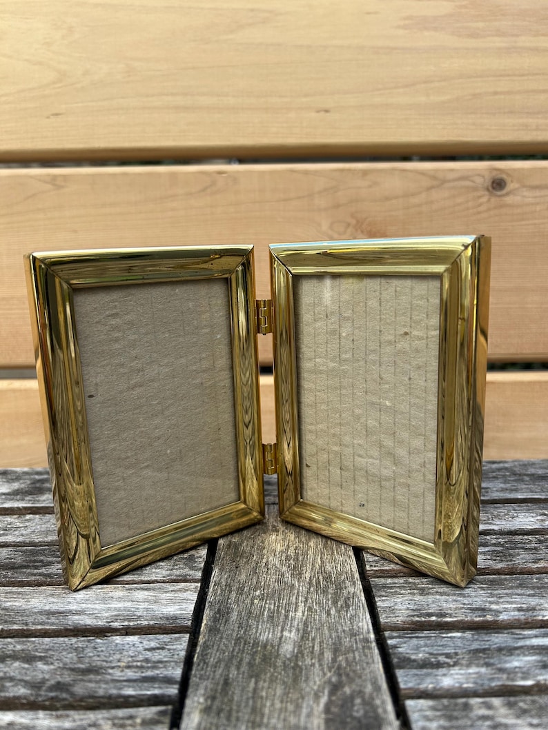 May include: A double-hinged picture frame constructed from polished gold-tone metal. The frame features two rectangular openings connected by a central metal hinge, allowing it to stand upright in a folded position with light beige inserts.