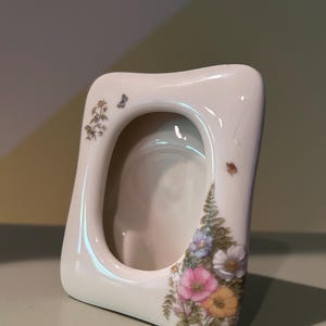 May include: A cream-colored ceramic picture frame with a smooth, rounded rectangular shape. The frame features a delicate floral pattern with pink, yellow, and white flowers, green fern leaves, and a small butterfly on the upper left side.