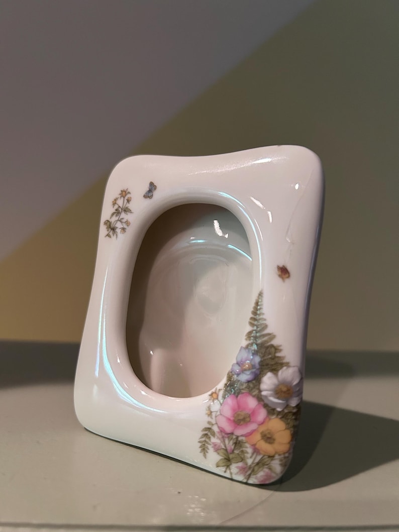 May include: A cream-colored ceramic picture frame with a smooth, rounded rectangular shape. The frame features a delicate floral pattern with pink, yellow, and white flowers, green fern leaves, and a small butterfly on the upper left side.