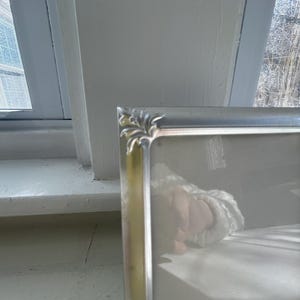 May include: A rectangular silver-toned picture frame featuring a smooth metallic finish and decorative corner accents with a subtle leaf-like embossed pattern. The frame includes a clear glass front panel and a clean, simple border design.