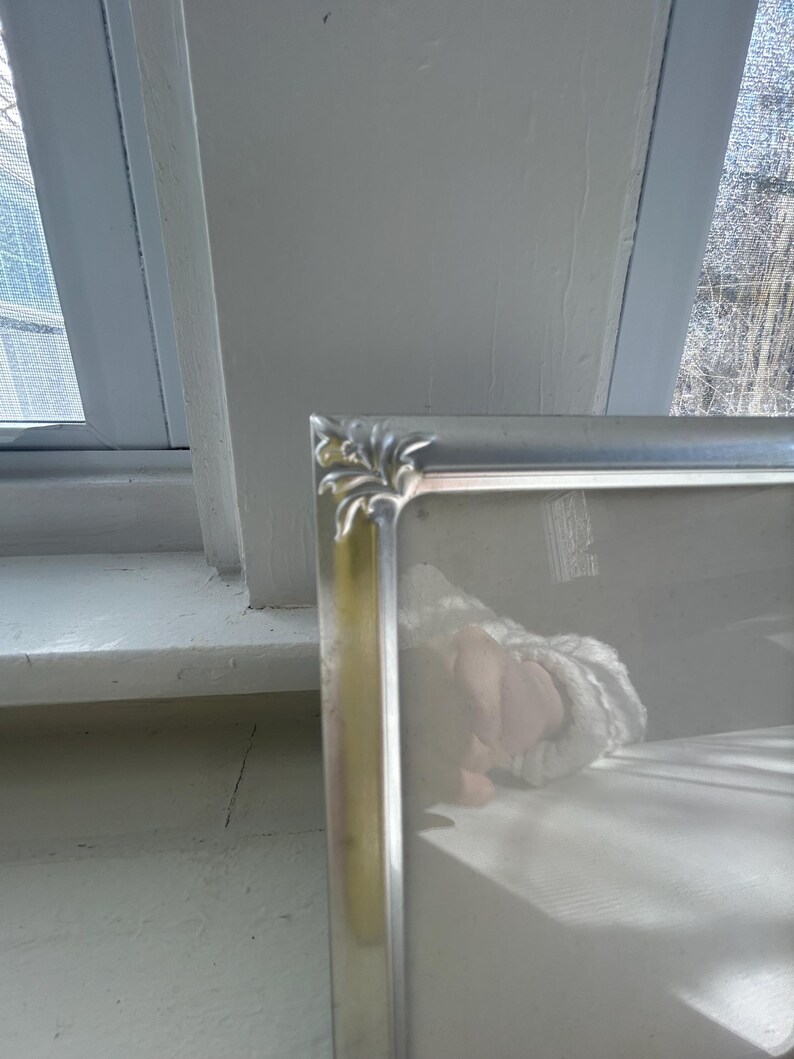 May include: A rectangular silver-toned picture frame featuring a smooth metallic finish and decorative corner accents with a subtle leaf-like embossed pattern. The frame includes a clear glass front panel and a clean, simple border design.