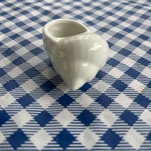 May include: A small, white ceramic creamer with a rounded body and a spout. The creamer is shaped like a small shoe or slipper. The background is a blue and white checkered pattern.