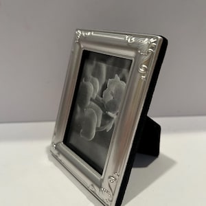 May include: A small rectangular picture frame with a metallic silver finish and ornate embossed detailing along the edges. The frame features a black velvet backing and a stand, holding a black and white photograph of a floral botanical subject.