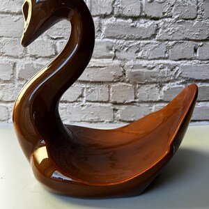 May include: A brown ceramic swan-shaped dish with a glossy finish. The swan has a long, curved neck and a wide, open body, suitable for decorative or functional use. The dish is set against a white and gray brick wall.