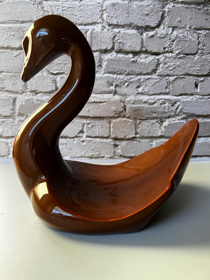 May include: A brown ceramic swan-shaped dish with a glossy finish. The swan has a long, curved neck and a wide, open body, suitable for decorative or functional use. The dish is set against a white and gray brick wall.