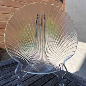 May include: A clear glass seashell-shaped plate with a textured surface, displayed on a clear acrylic stand. The plate has a scalloped edge and radial lines emanating from the center. The stand has three legs.